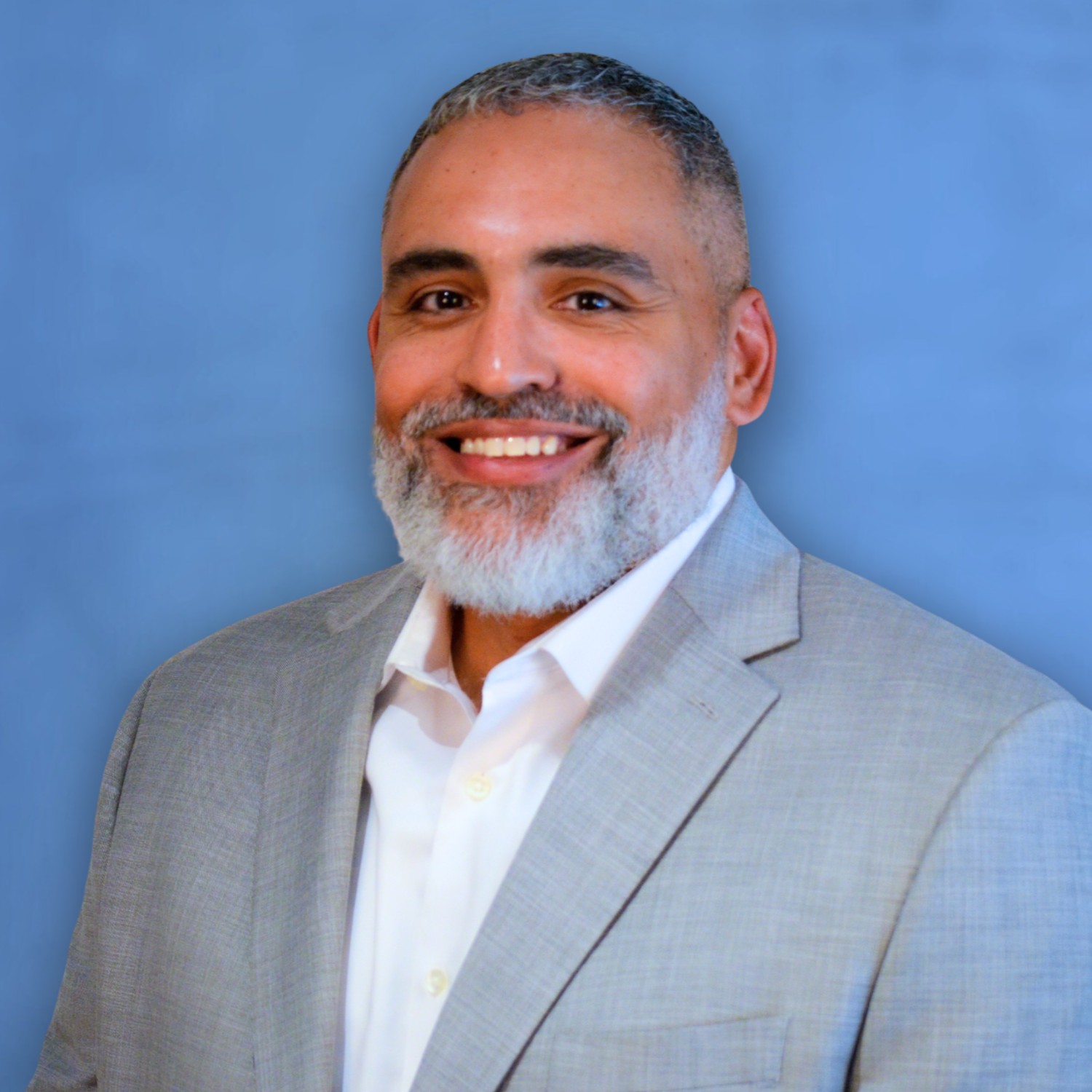 Professional head-and-shoulders portrait of a person wearing a light gray blazer over a white button-down shirt, posed against a solid blue background, facing the camera.