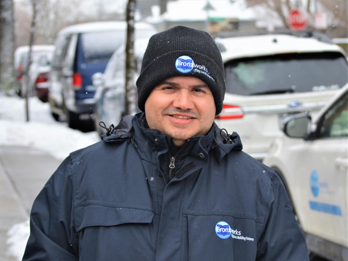 A male BronxWorks staff standing in the side walk on a snowy day smiling at the camera