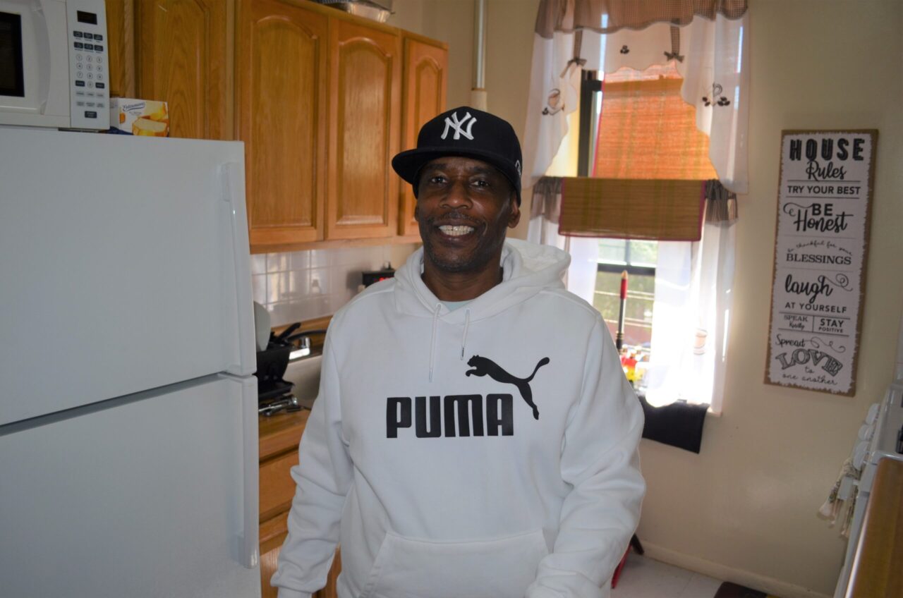 A man standing in the middle of a kitchen smiling at the camera wearing a yankees hat and puma hoodie