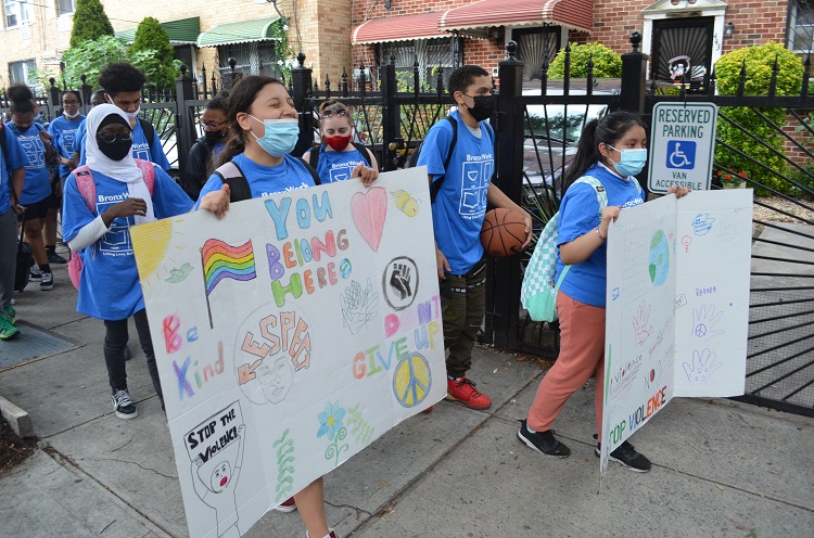 Students in blue BronxWorks shirts march along the street carrying signs with messages of peace, including "You Belong Here" and "Be Kind". 
