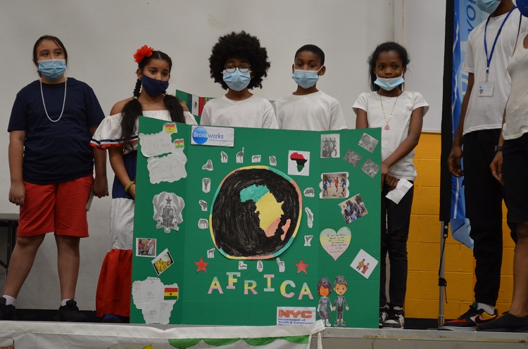 Five children stand behind a green posterboard. There is a cutout of Africa and other smaller pictures.