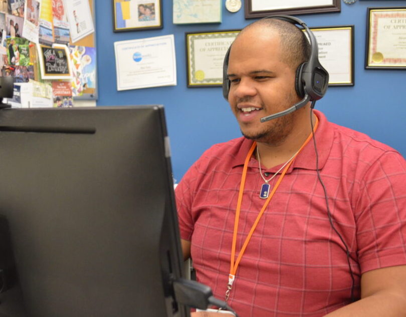 A man in a red shirt wearing a headset speaks to a virtual training session on his computer.