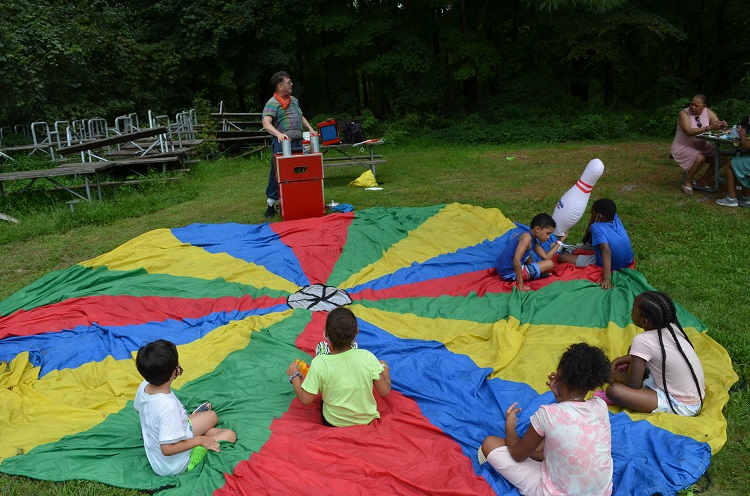 Young children sit on a parachute while being entertained by a clown.