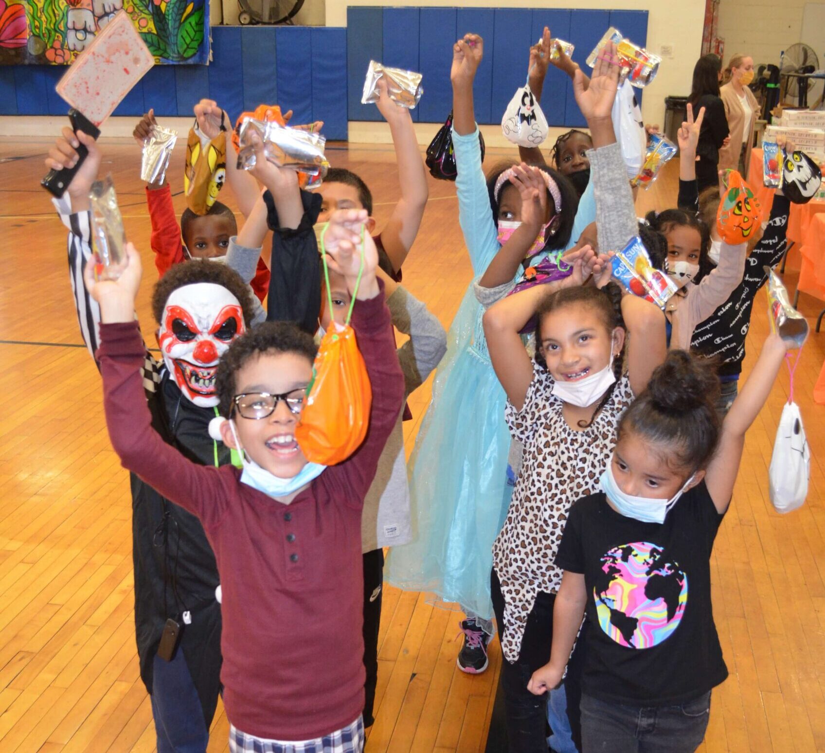 A group of children hold their hands up in the air and cheer. They are holding Halloween bags.