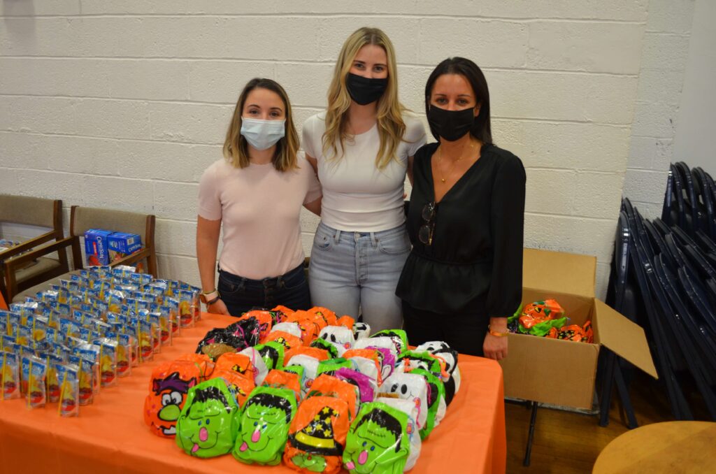 Three women wearing masks stand behind a table with goody bags decorated with Halloween creatures.