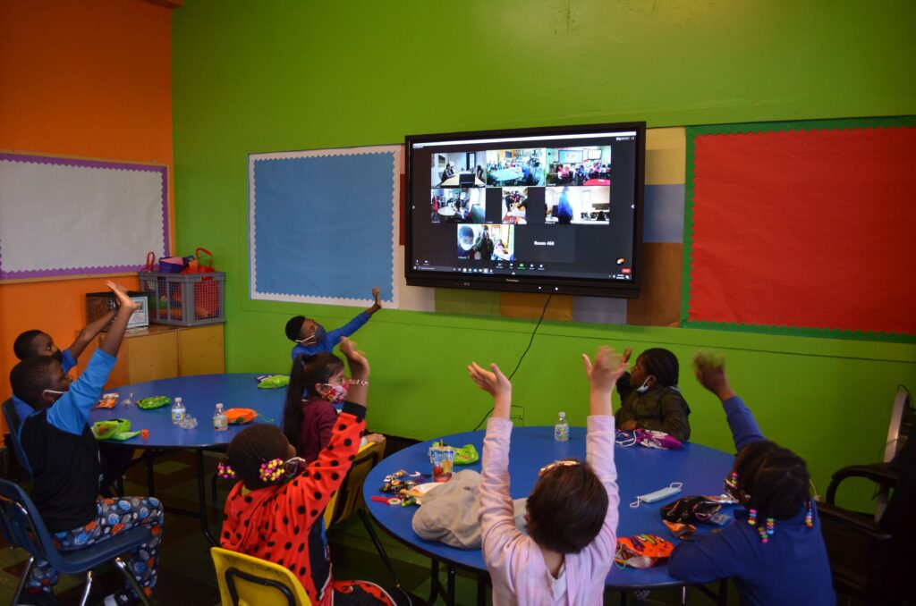 Groups of children wave at a monitor showing a Zoom meeting.