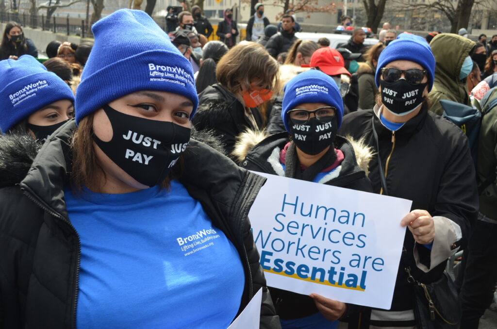 BronxWorks staff wearing BronxWorks hats and Just Pay masks, holding a sign saying Human Services Workers are Essential