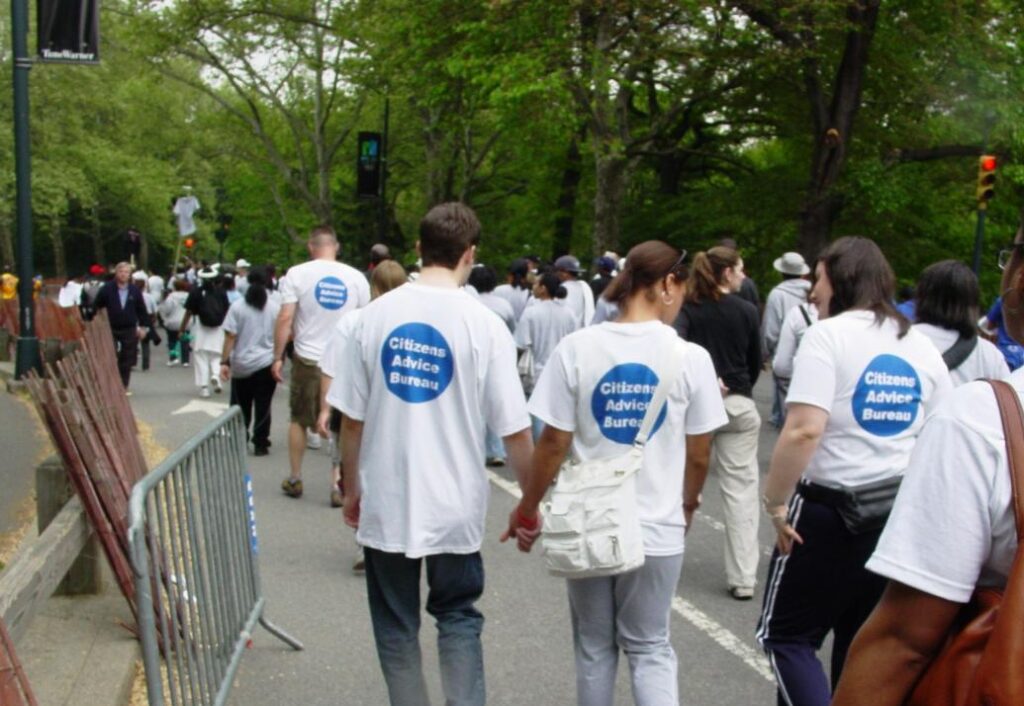 A group of people walking in Central Park in 2005 at the AIDS Walk NEw York. The shirts are emblazoned with a blue circle containing the text "Citizens Advice Bureau".