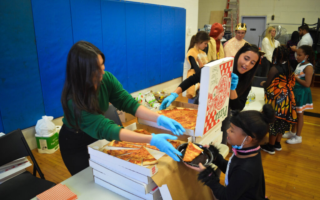 Two women hand out pizza to a child dressed as a cat.