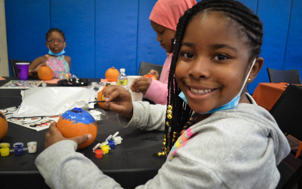 A girl smiles at the camera while painting a pumpkin.