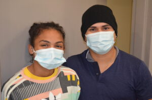 Two people wearing face masks and smiling. The person on the right is wearing a knit cap.