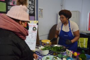 A woman in a blue apron shows another person how to make a kale and apple salad at an event.