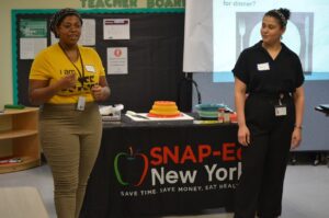 Two women speaking to a crowd. There is a black tablecloth printed with the SNAP-Ed NY logo on it.