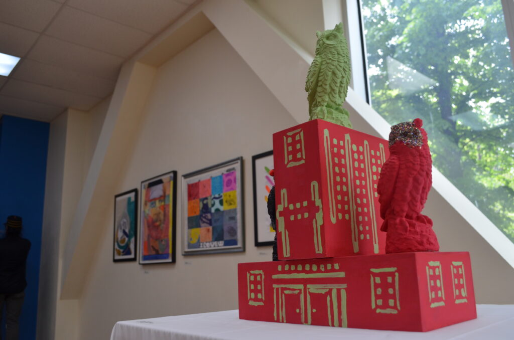 “Safe House” by Manny R. on display at the Boricua College Art Gallery. The sculpture looks like a red building, with gold doors and windows painted on. The building is adorned with three owls in black, green, and red.