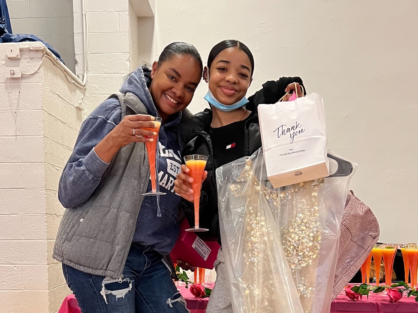 A mother and daughter hold up a prom dress, a gift bag, and a mocktail.