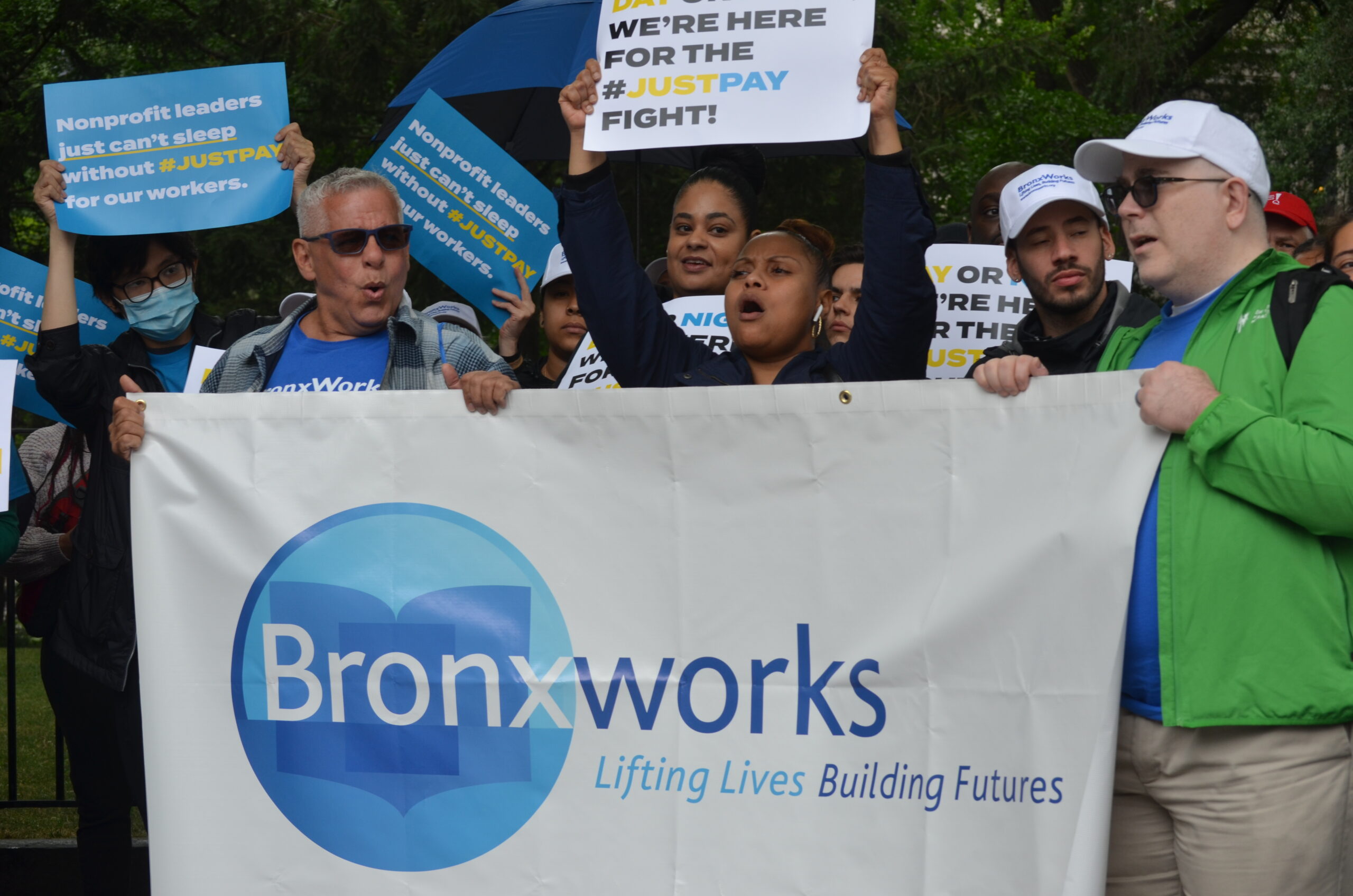 Multiple people rallying holding signs. There is a large banner with the BronxWorks logo. One woman holds a sign that says "We're Here for the #JustPay Fight"