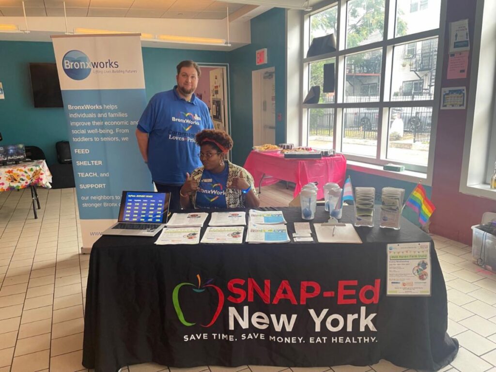 BronxWorks staff members at a BronxWorks Pride Event. There is a BronxWorks sign behind them. The tablecloth has a SAP-Ed New York logo on it.