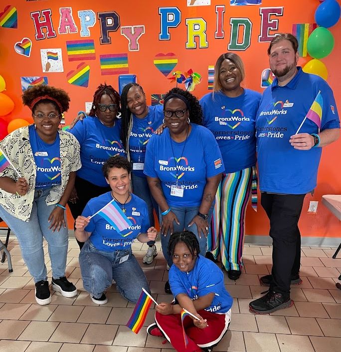 Staff wearing shirts that say "BronxWorks Cornerstones Loves Pride". Many are holding pride flags. There is a sign behind them reading "Happy Pride".