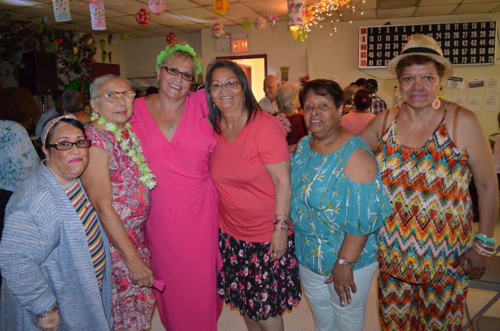 Women stand in a room decorated like a party. Many have leis or flower crowns on.