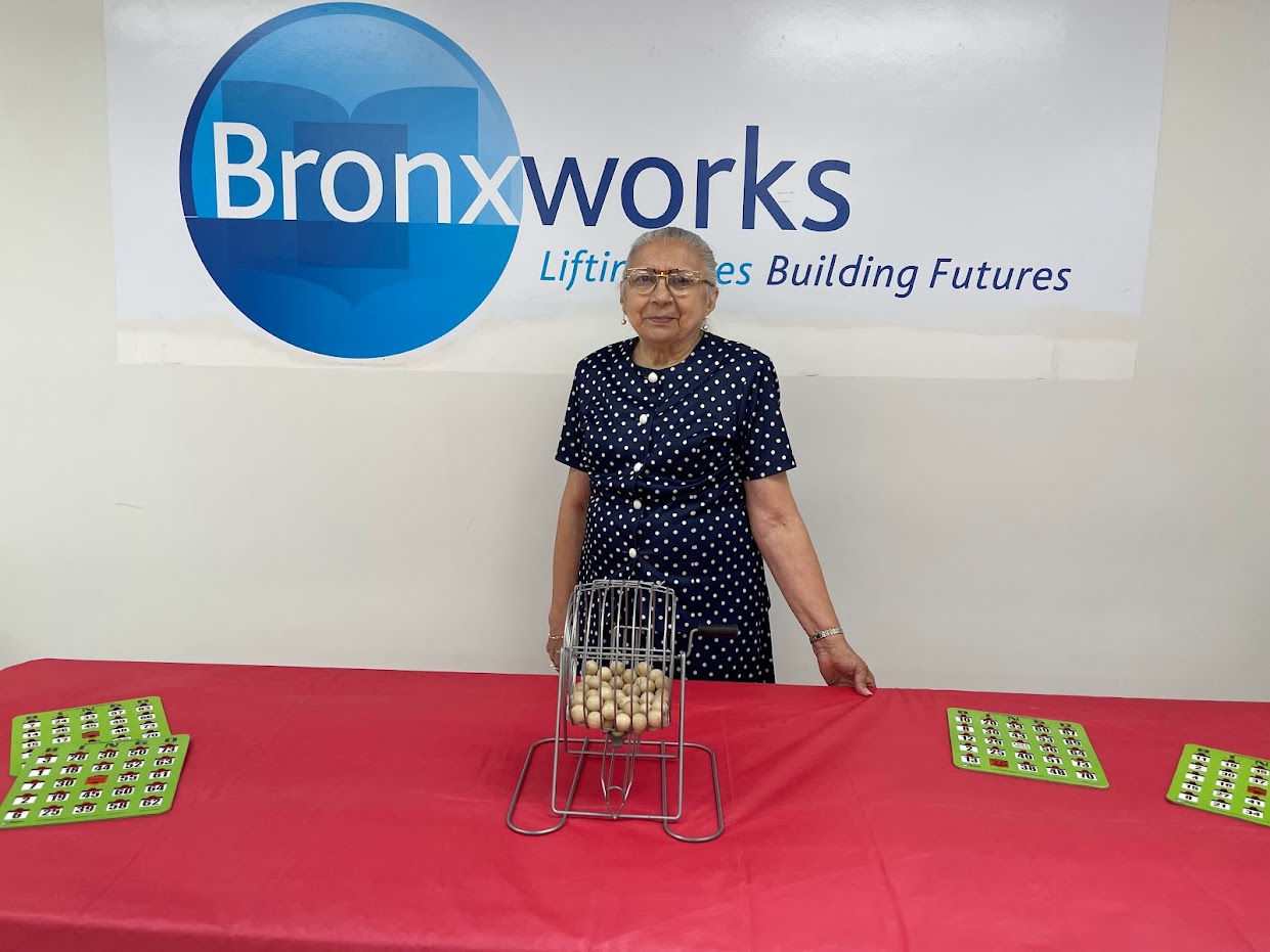 A woman stands behind a Bingo cage and in front of the BronxWorks logo. She is smiling.