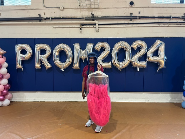 A Girl holds a pink prom dress in front of silver balloons that spell out PROM 2024