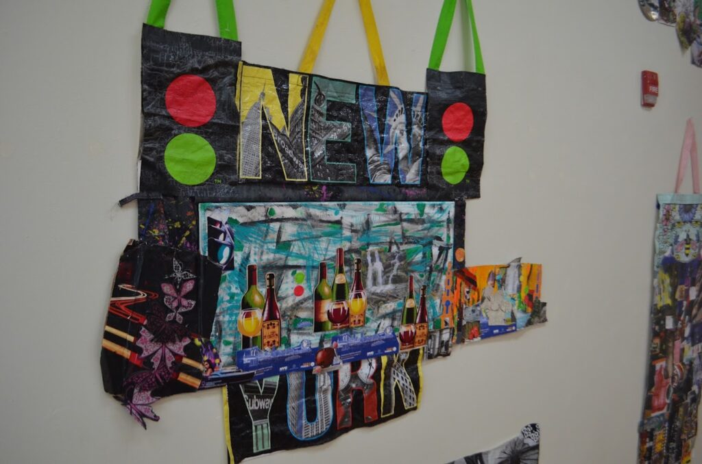 Artwork on a wall. The piece is a cut-up reusable shopping bag. The bag prominently says NEW YORK in block letters. The letters are each filled in with images from around New York City. The rest of the piece shows a collage of various other images, including wine bottles and other floral designs.