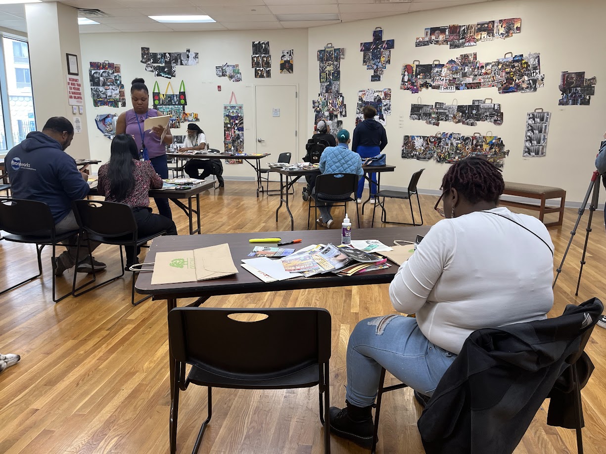 Many people seated in an art gallery. There is artwork on the walls. The people in the gallery are seated at tables and all working on various art projects.