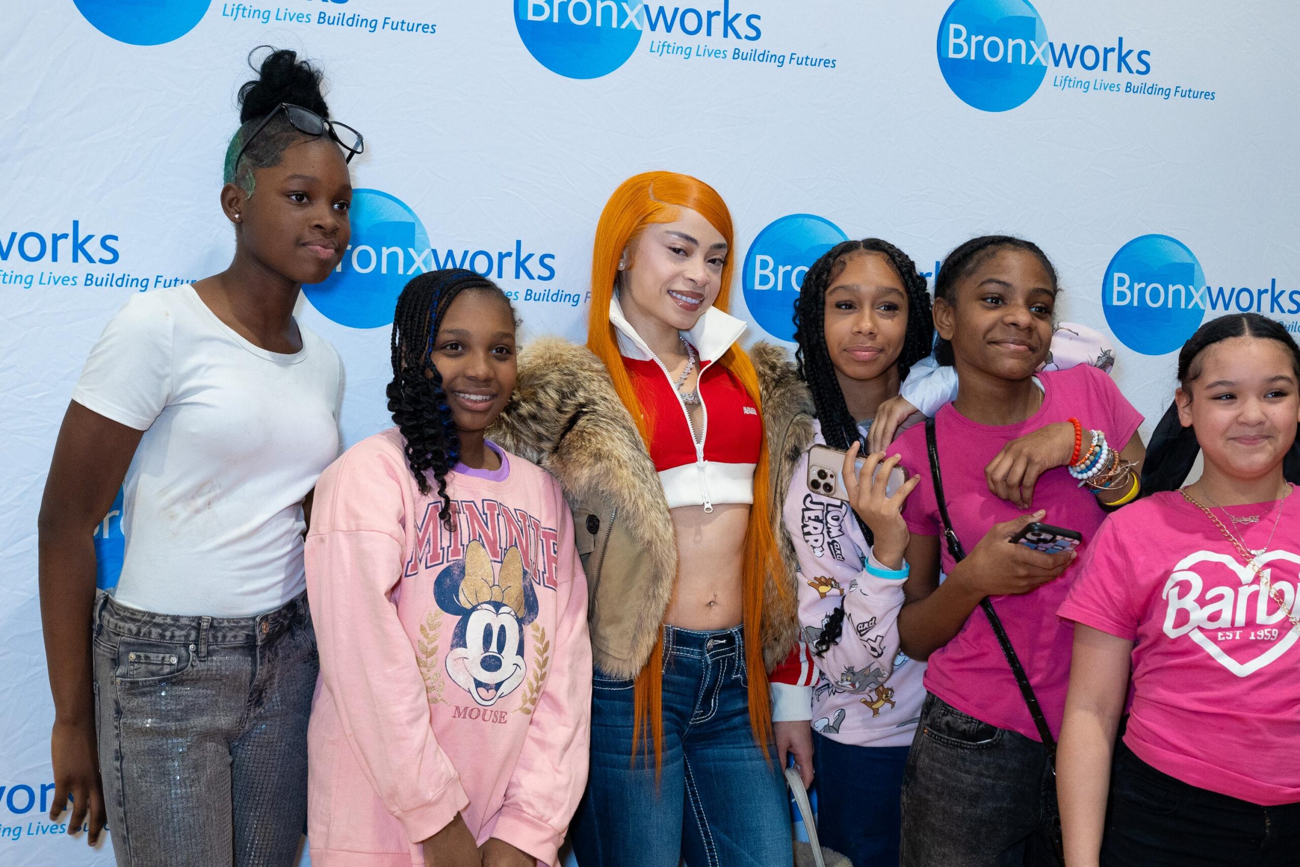 Girls stand in front of a BronxWorks step & repeat smiling. Rapper Ice Spice stands in the center.