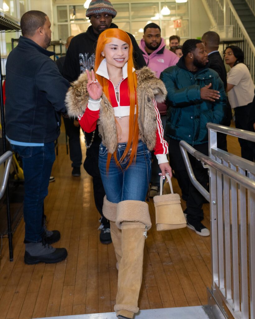 Rapper Ice Spice waves and smiles as she walks up a metal ramp. Many people stand behind her talking.