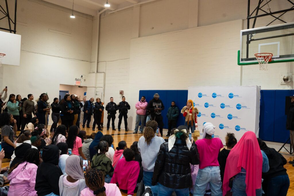 Many people stand in a gymnasium. Rapper Ice Spice stands in front of a crowd of girls and speaks into a microphone.