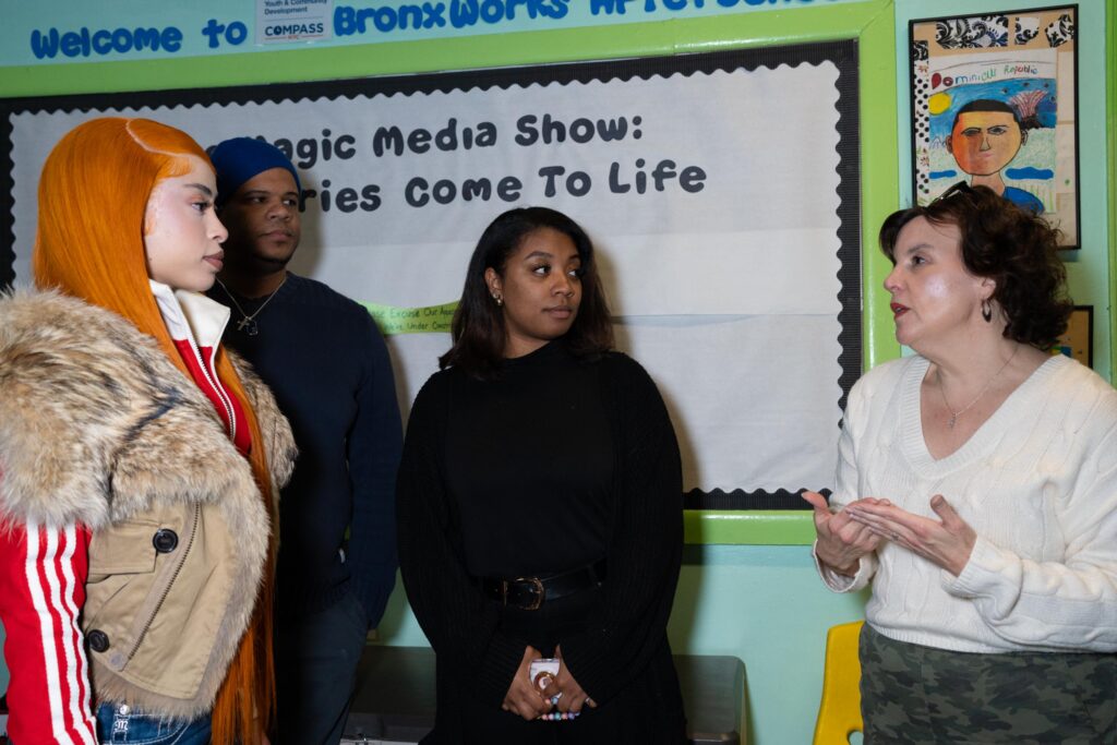 Rapper Ice Spice and others look on as a BronxWorks staff member talks about BronxWorks. They are standing in a hallway with art and signs on the walls.