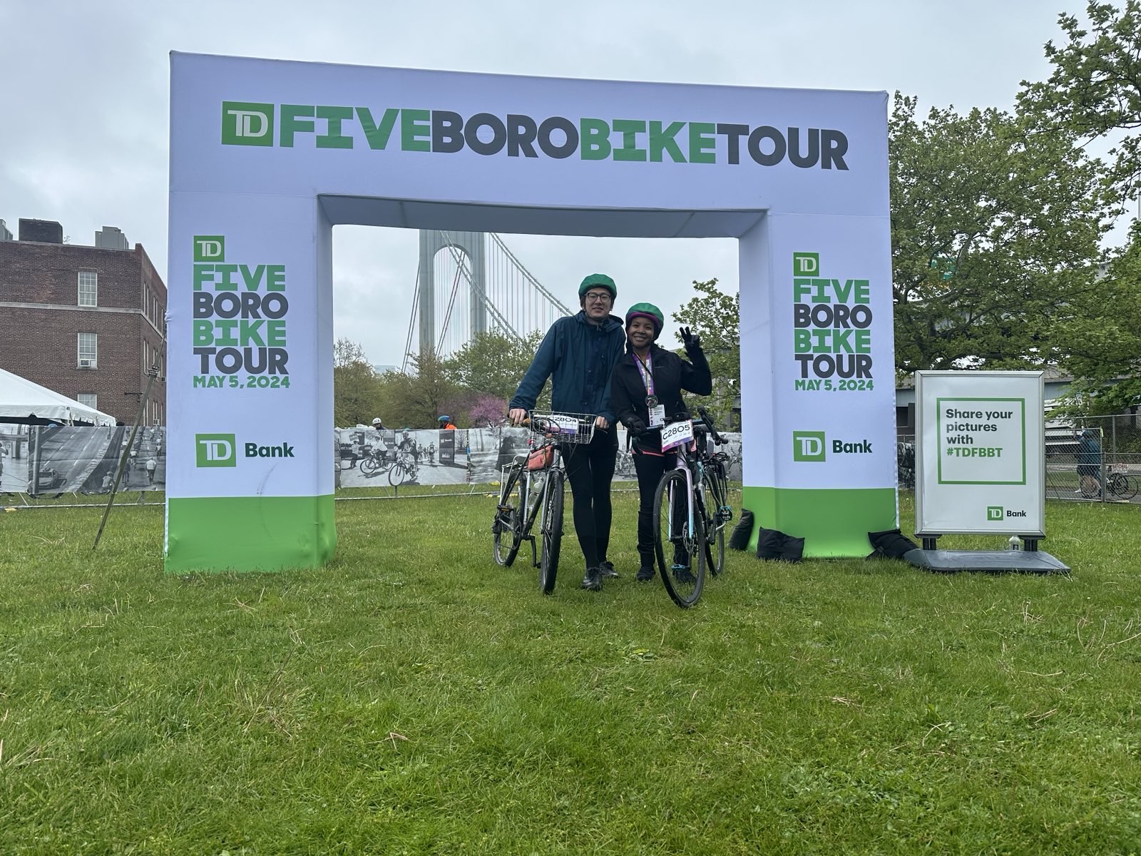 Two cyclists stand under an archway with the TD Five Boro Bike Tour logo on it.