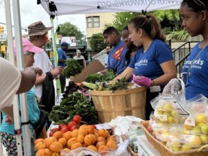 There are BronxWorks staff on the right behind a display of fruits and vegetables. On the right are people shopping for those items.