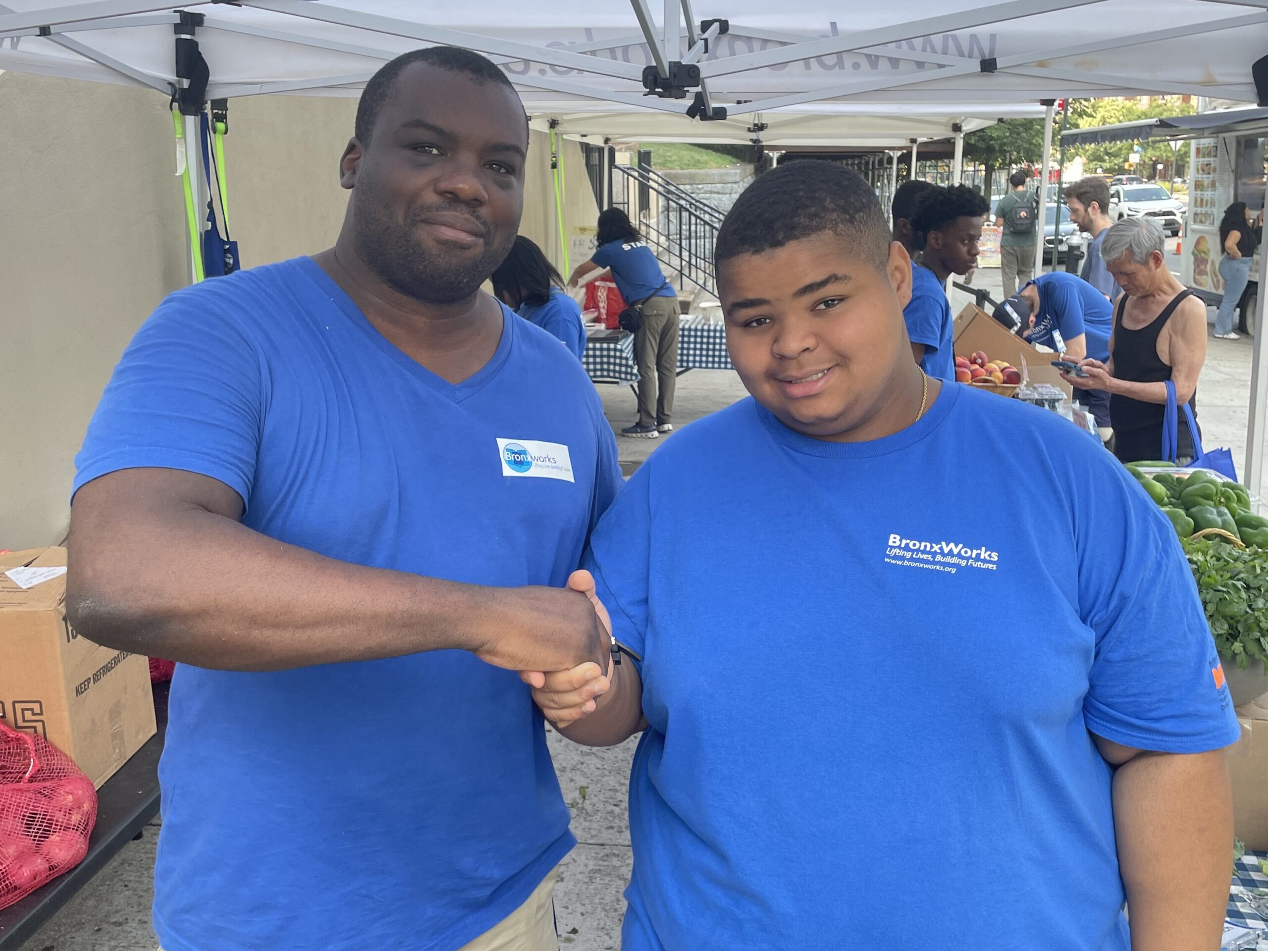 Two men shake hands. They are both wearing blue BronxWorks t-shirts. There is a farm stand behind them.