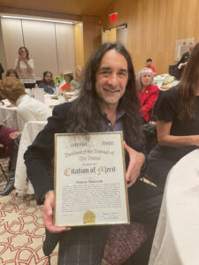 A man with long black and gray hair holds a Citation of Merit