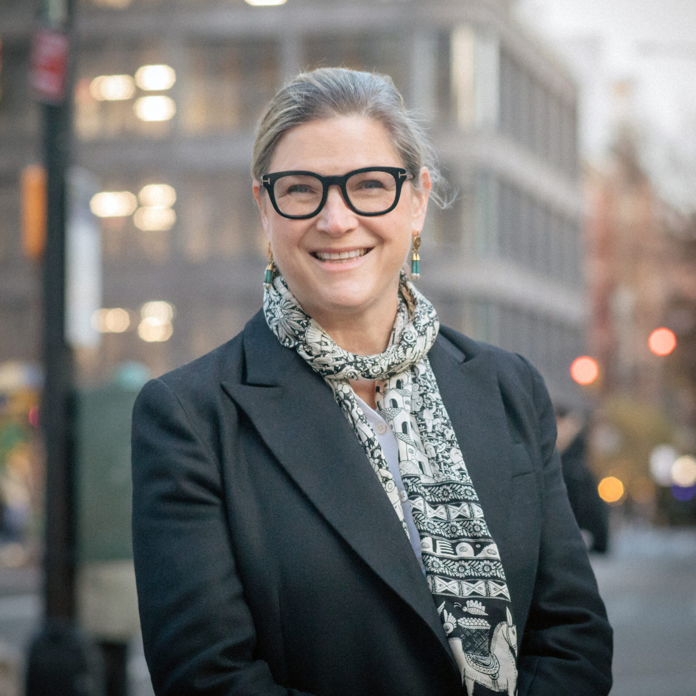 Woman wearing glasses and a dark blazer stands on a city street with buildings and traffic lights in the background.