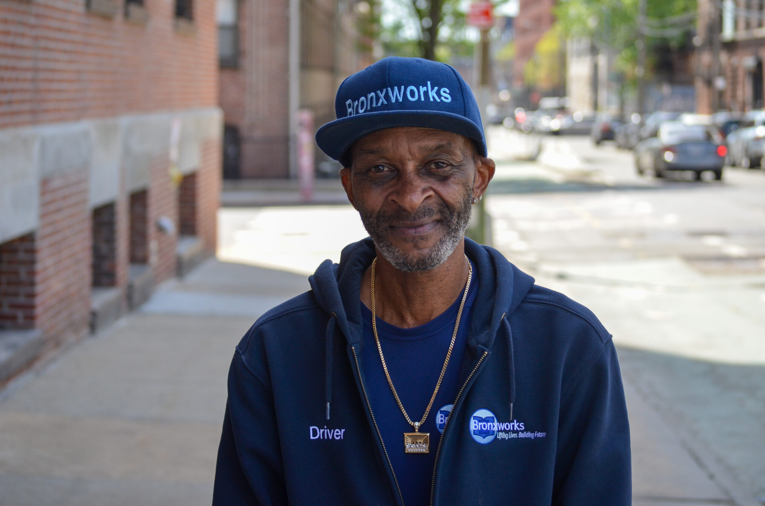A picture of a smiling man in a BronxWorks hat and jacket standing outside on the sidewalk next to a building.