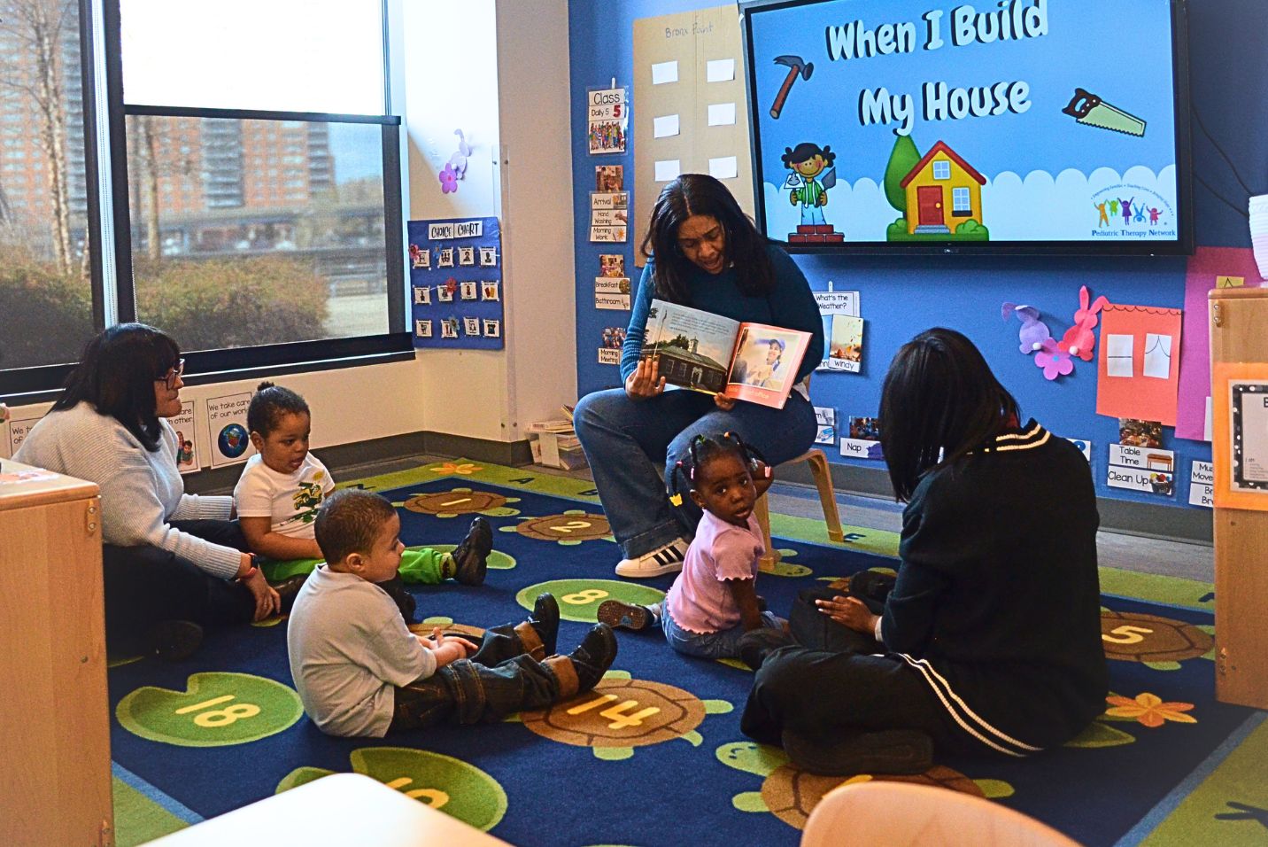 An adult reads a picture book to a small group of children seated on a colorful classroom rug, while two other adults sit nearby. The classroom includes a wall display reading “When I Build My House,” shelves, windows, and educational decorations.