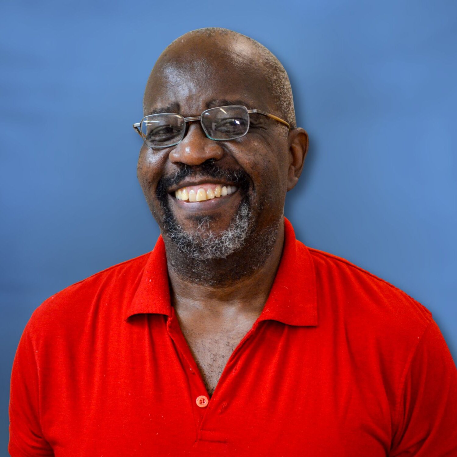 Portrait of a smiling person wearing glasses and a red collared shirt, facing the camera, standing against a plain blue background.