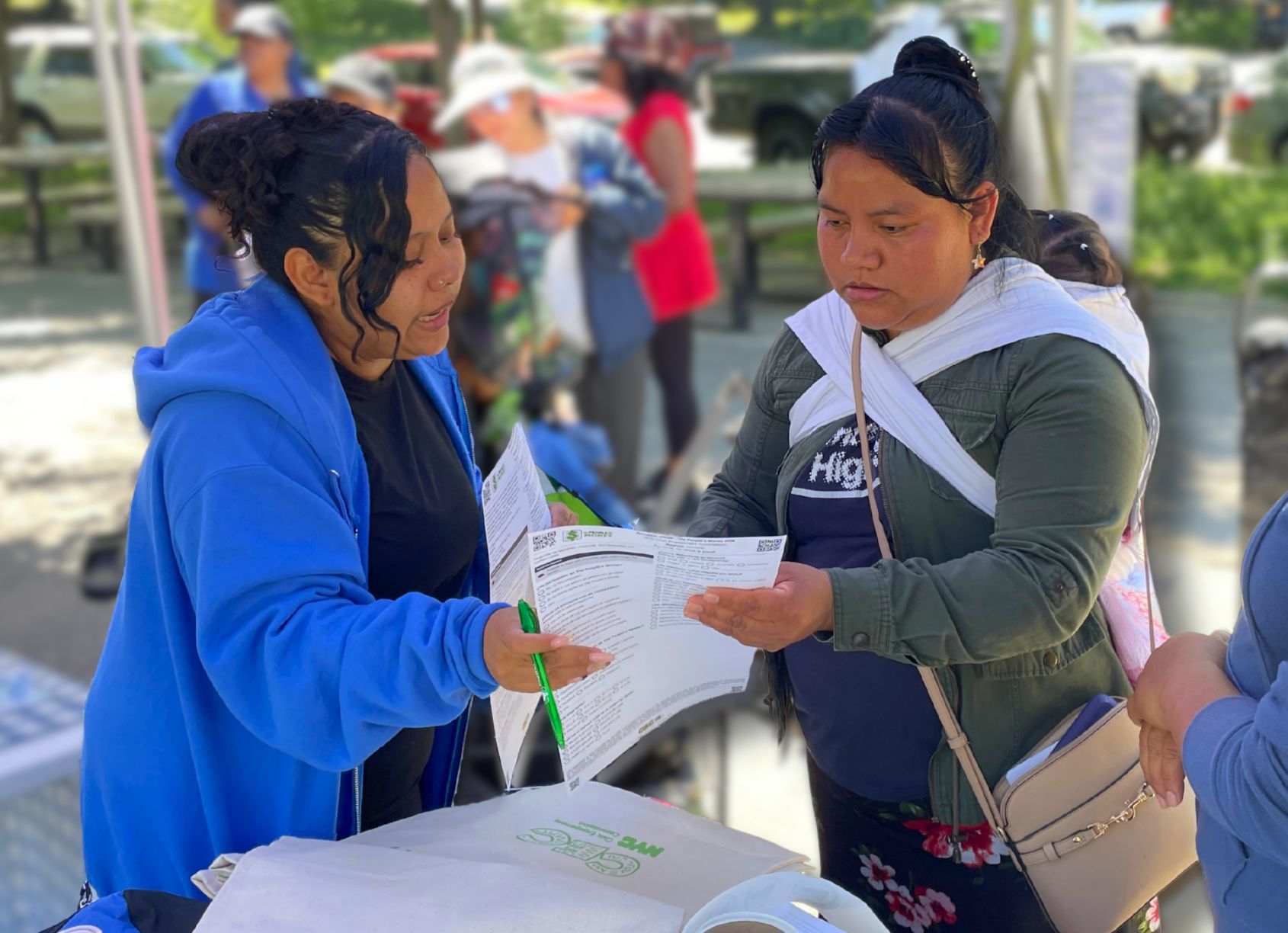 A person in a blue hoodie helps another person review and complete paperwork at an outdoor community resource event. The two stand at a table with forms and tote bags while several other attendees are visible in the background.