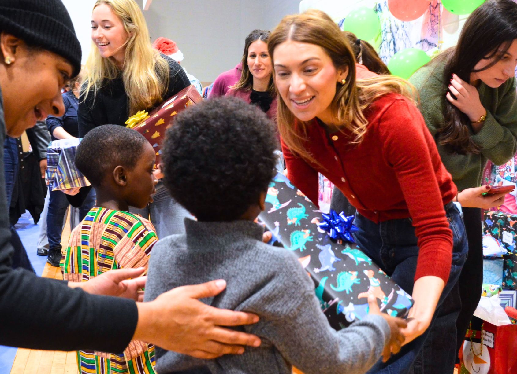 Volunteers hand wrapped gifts to children at a holiday celebration. Several volunteers and families gather in a decorated room with balloons and colorful gift bags as a child receives a large wrapped present.