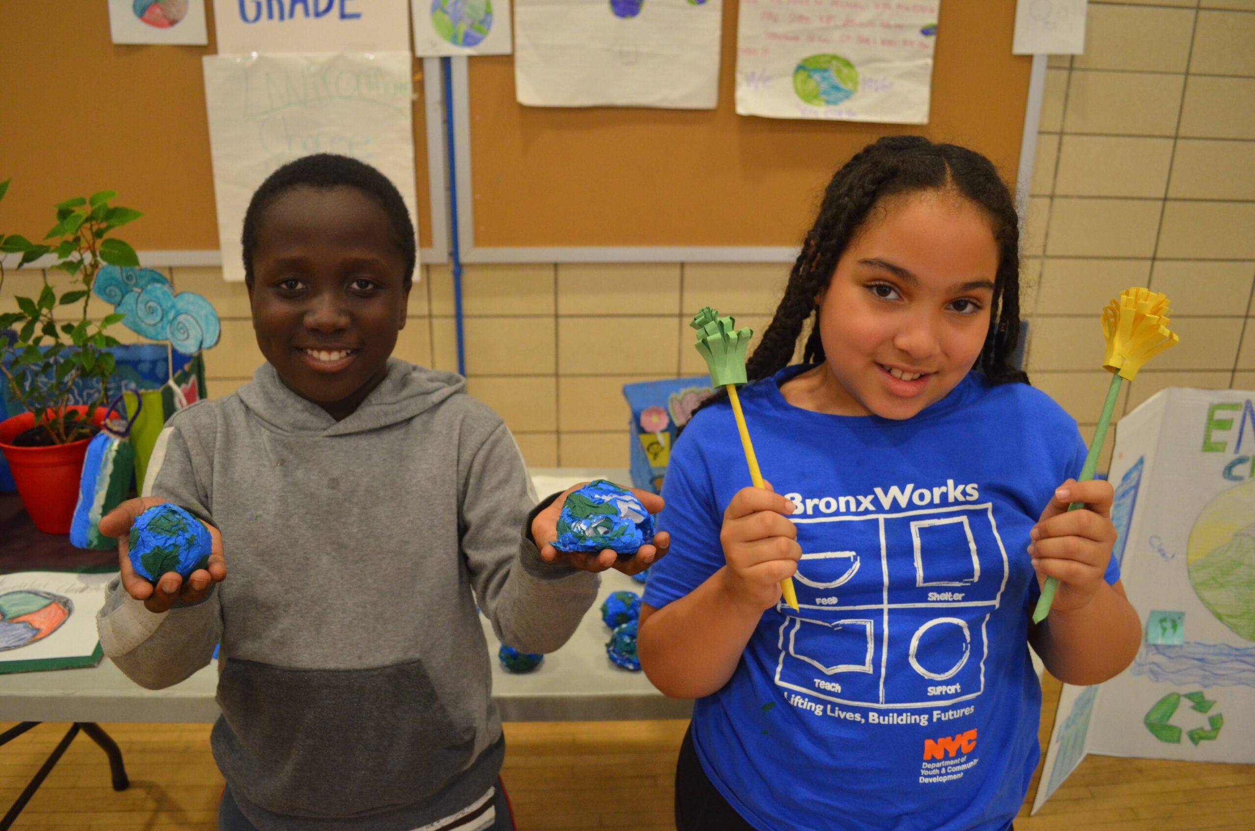 Two children stand behind a display table, each holding handmade Earth-themed projects. One child holds painted globe models, while the other holds craft flowers made from colored paper. Posters and artwork about environmental themes and Earth Day are displayed on bulletin boards and easels in the background, creating a classroom or school project showcase setting.